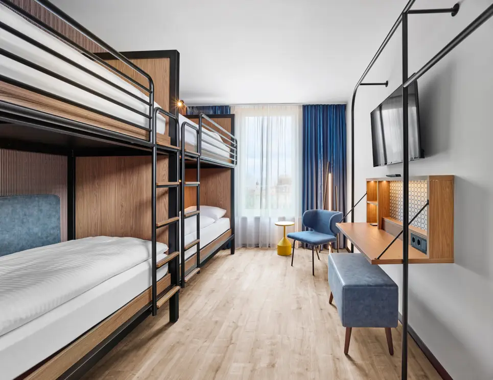 Bunk beds in the hotel room. There is a chair with a table in the corner. A television hangs on the wall to the right, underneath is an open desk and a wide stool.