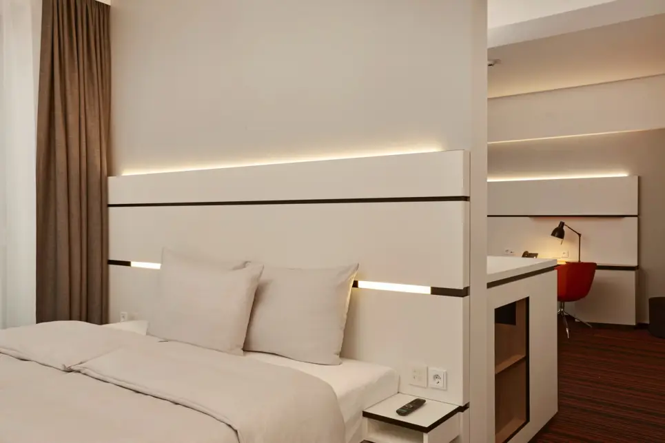 Schlichtes, modern Hotelzimmer mit beleuchtetem Kopfteil. Im Zimmer hinter dem Bett ist vage ein Schreibtisch zu erkennen.